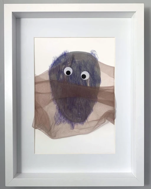 Whimsical mixed-media artwork: furry blue oval face with googly eyes peering through a brown semi-sheer fabric, framed in white.