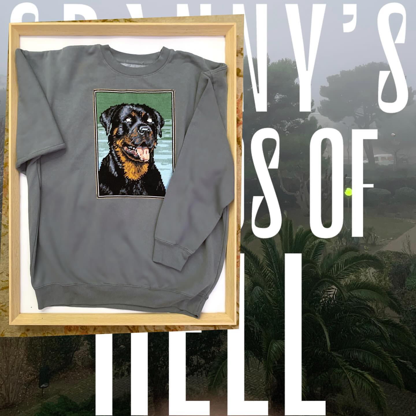 Grey sweatshirt featuring a Rottweiler graphic, showcased in a wooden frame against a backdrop of blurred text and landscape.