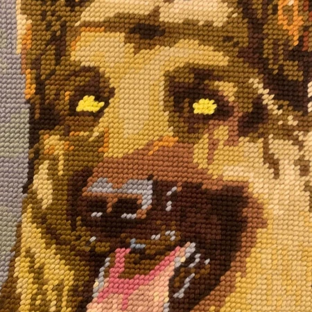 Close-up of a needlepoint tapestry featuring the face of a brown and tan dog with its mouth open, tongue showing and yellow eyes.