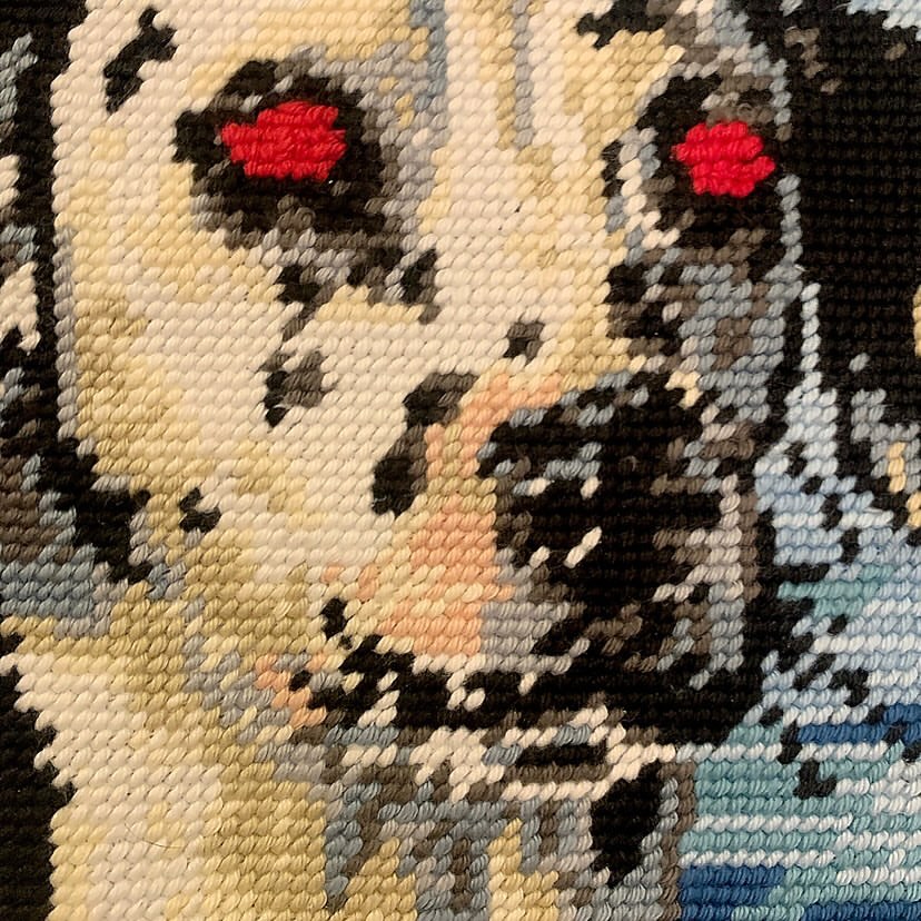 Close-up needlepoint image of a dog's face with light fur and black spots, set against a textured, hand-crafted background.