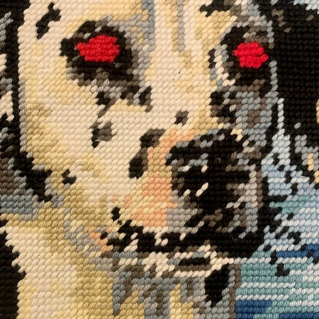 Close-up needlepoint image of a dog's face with light fur and black spots, set against a textured, hand-crafted background.