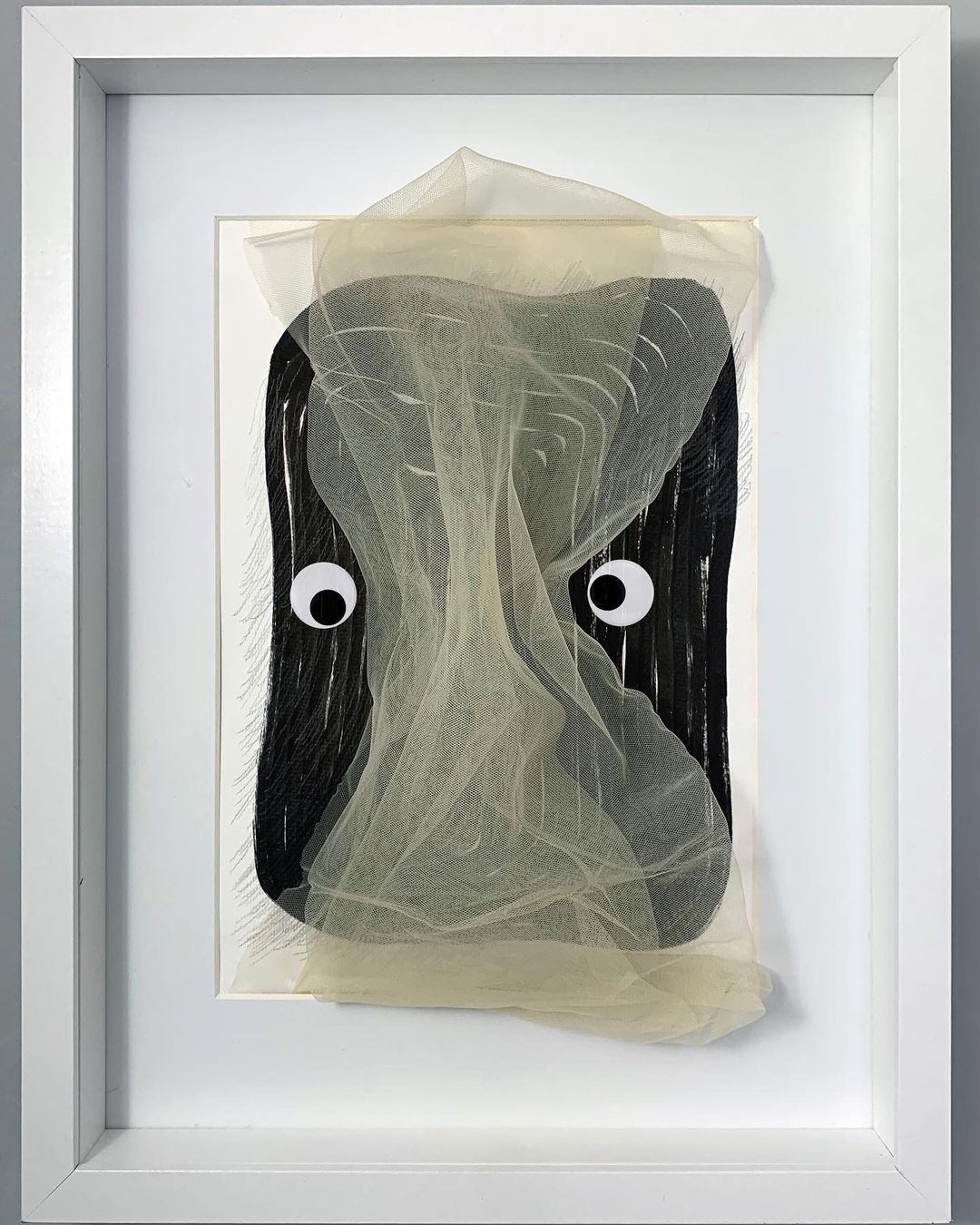Whimsical wall art featuring an abstract, veiled face with googly eyes within a clean white frame, creating a playful, surreal aesthetic.