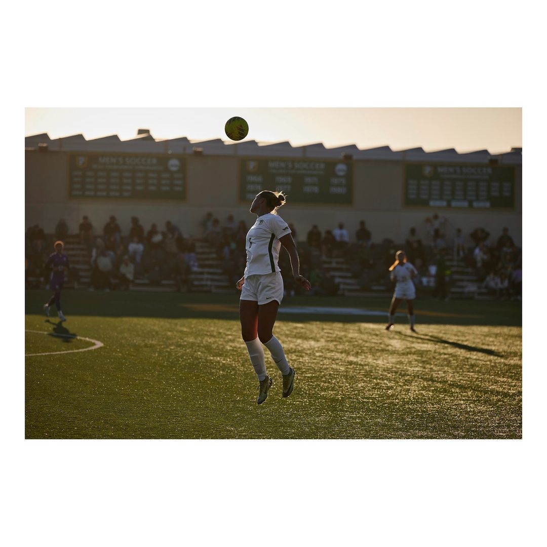 Female soccer player jumping to head a soccer ball on a green field during a game with spectators and banners in the background.
