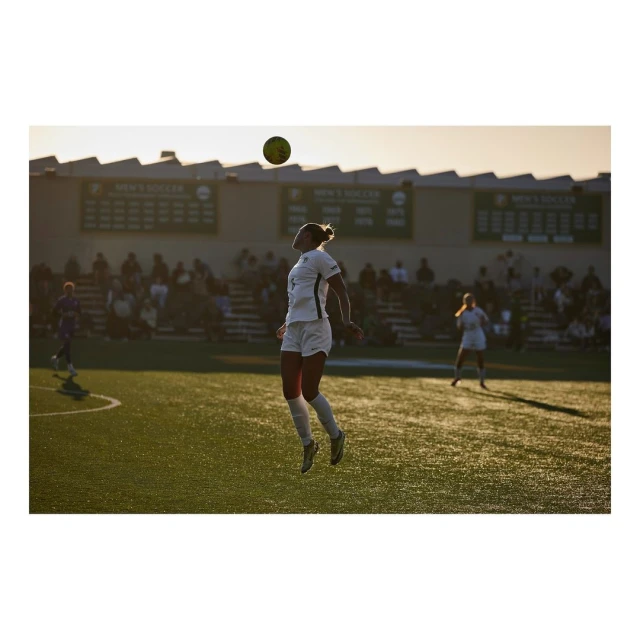 Female soccer player jumping to head a soccer ball on a green field during a game with spectators and banners in the background.