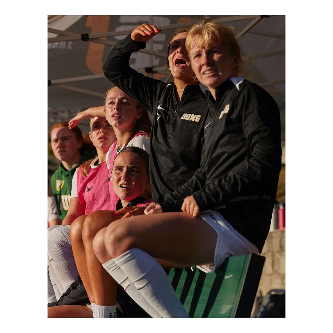 Women's soccer team intently watches the game from the sidelines on a sunny day, wearing uniforms and team jackets, seated on the bench.