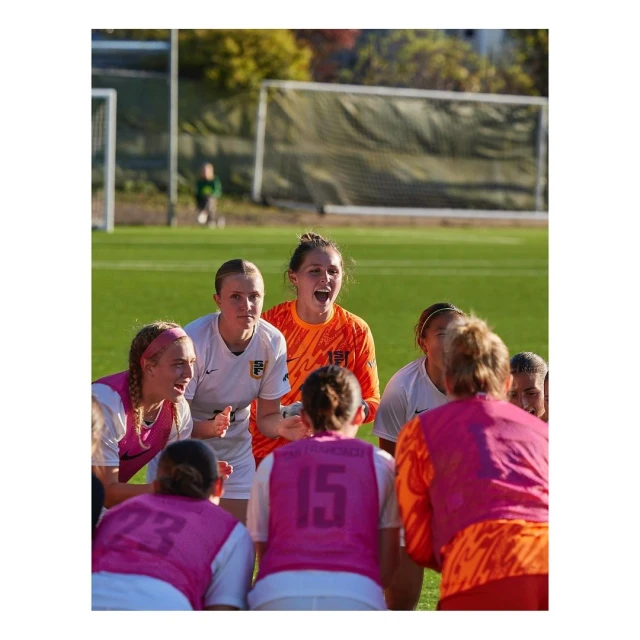 Outdoor shot of a female soccer team in uniform gathered on a green sports field, with one player in orange vocally leading the team.
