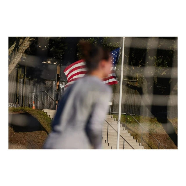 A blurry figure walks past an American flag on a pole, viewed through the netting of a sports goal on a sunny day.