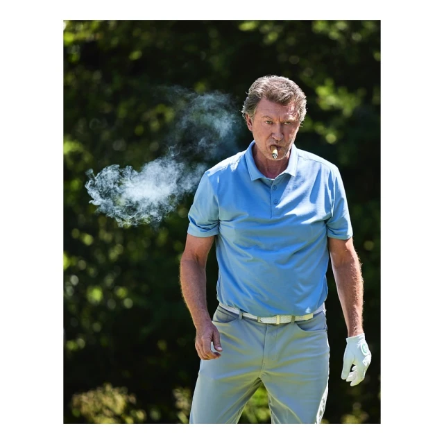 Mature man in blue polo shirt outdoors smoking cigar, set against a blurred green background of trees and foliage on a sunny day.