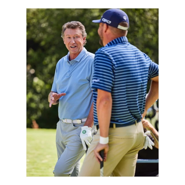 Two adult men in golf attire converse on a sunny green golf course, one wearing a blue polo and khaki pants, the other wearing a blue striped polo.