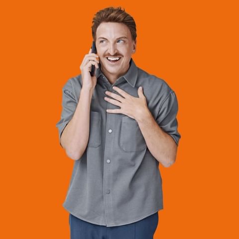 Adult male with a mustache in gray button-down shirt talking on cell phone with an orange background in an indoor photo studio.