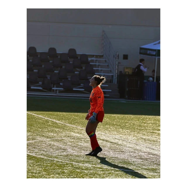 Female soccer player in uniform standing on a green field with stadium seating visible in the background, captured in side view.