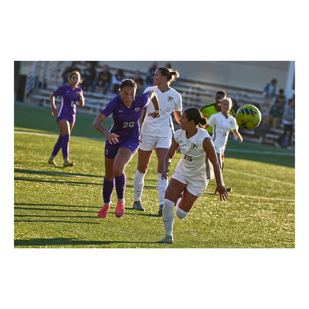 Female soccer players in action on a sunny green field wearing white and purple jerseys during an outdoor game on a sunny day.