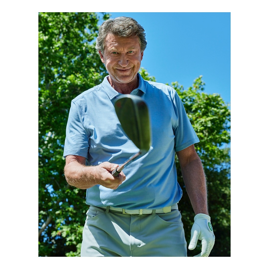 Adult male golfer holding a golf club on the green outdoors. The sky is blue, and trees are visible in the background.