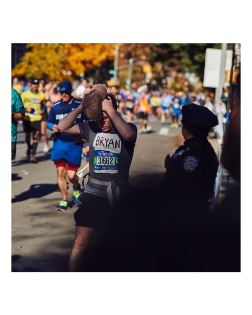 Marathon runner adjusts cap during outdoor race on city street with spectators and law enforcement presence. Event showcases fitness, sport.