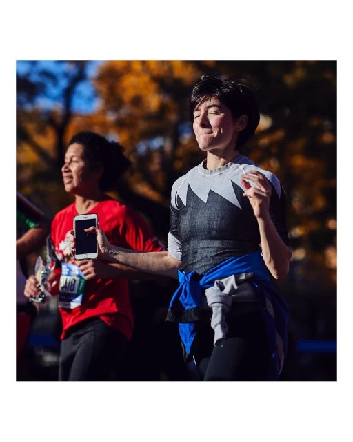 Two women participate in an outdoor marathon on a tree-lined street, with one holding a phone and the other wearing a race bib.