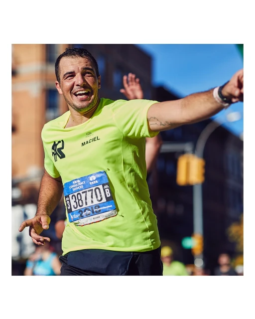 A man wearing a bright yellow shirt smiles during a marathon in an urban setting with a traffic light, buildings, and a blue sky.