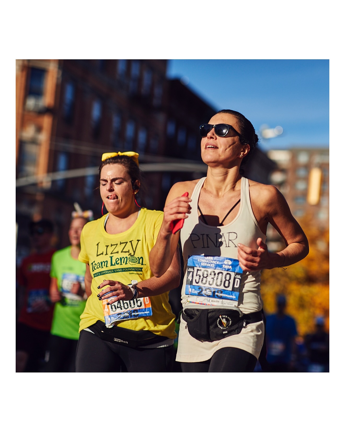 Two adult females running in a city marathon race on a sunny day with buildings in the background and blue sky overhead.