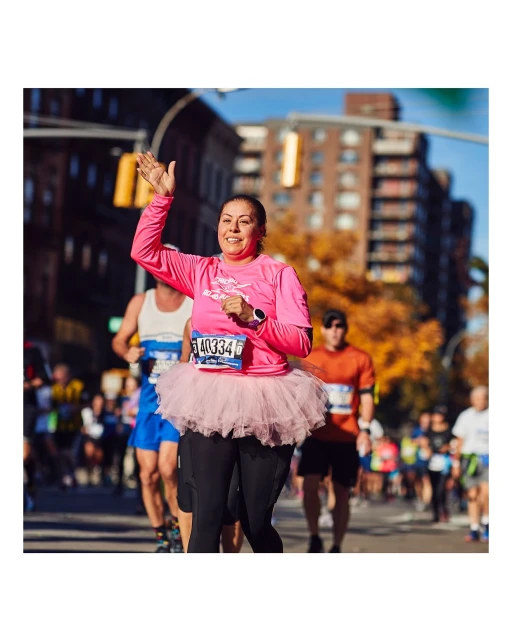Adult female runner in a pink shirt and pink tutu waves to the crowd during a marathon in an urban setting on a sunny day.