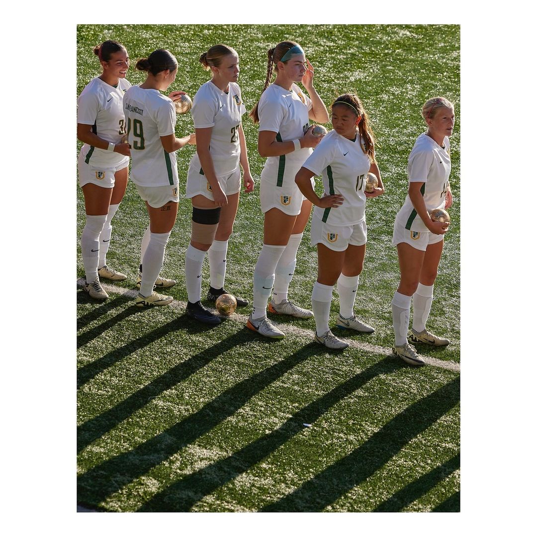 Group of young female soccer players in white and green uniforms standing on a green field with long shadows, holding soccer balls.