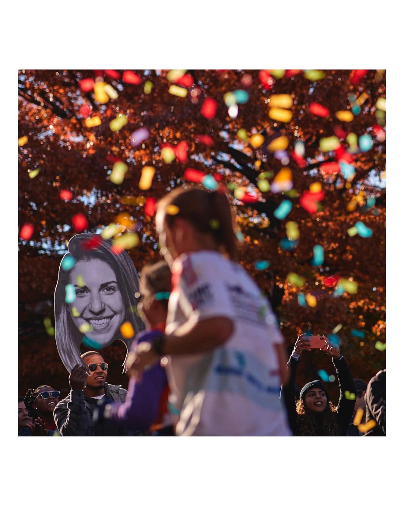 Crowd of people celebrating with confetti falling, someone holds a large face cutout in an outdoor setting, autumn leaves visible.