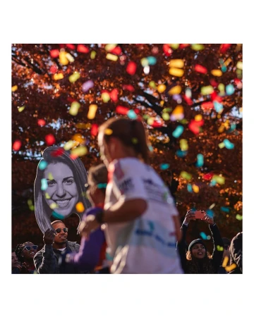Crowd of people celebrating with confetti falling, someone holds a large face cutout in an outdoor setting, autumn leaves visible.