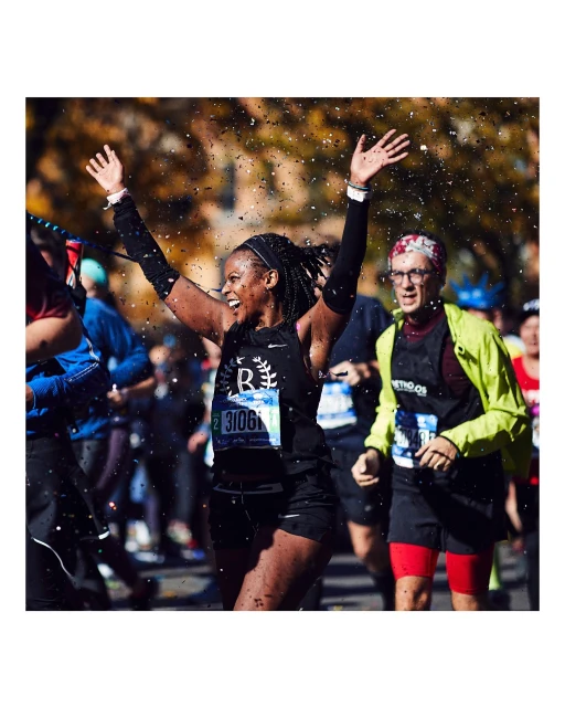 A woman runner with arms raised crossing the finish line with confetti, cheered on by other marathon runners during an outdoor race.