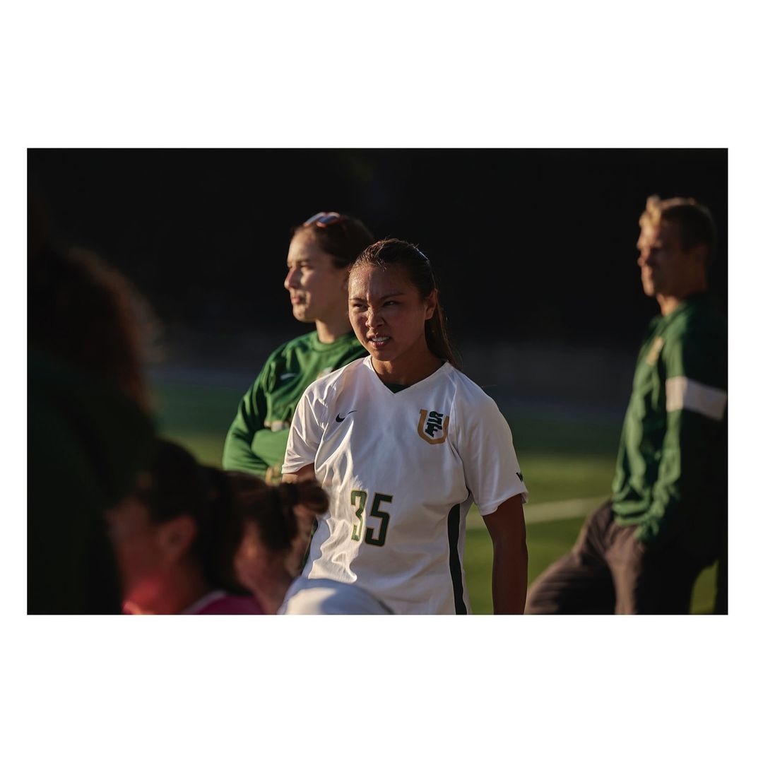 Female soccer player in white and green jersey on the field during a game, with blurred teammates in the background. 