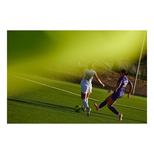 Two female soccer players compete on a green field under bright sunlight, one kicking the ball while the other defends. 