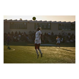 Female soccer player jumping to head a ball on a turf field during sunset with spectators in the background.