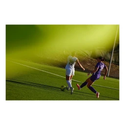 Two female soccer players competing on a green turf field with a bright green light leak overlaying the upper portion of the frame.