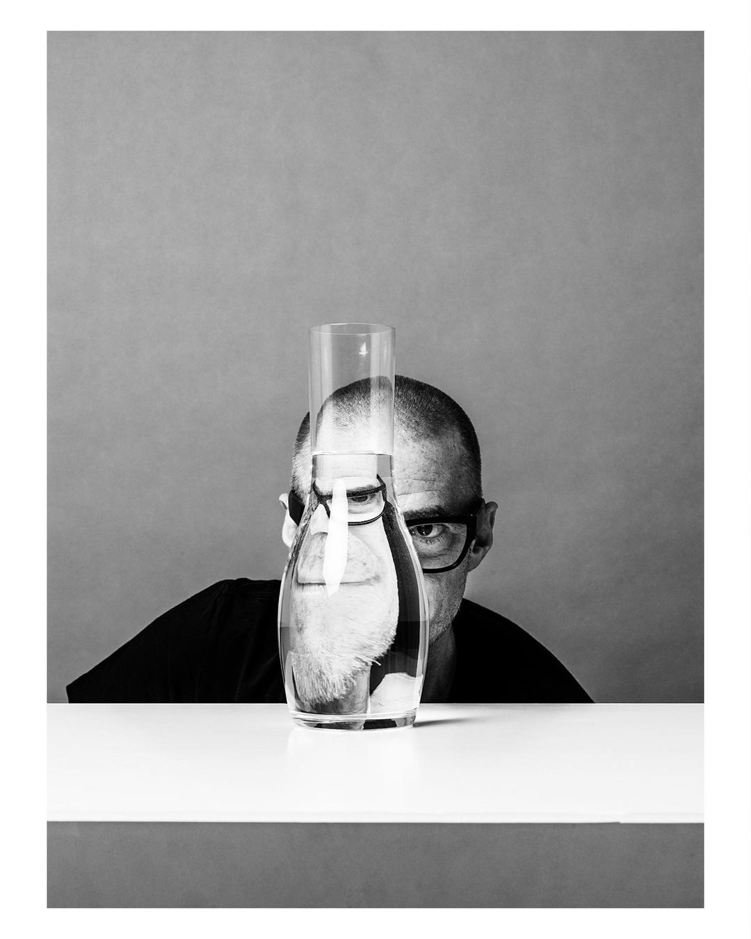 Monochrome portrait of a bald adult man with glasses peering over a table, a water carafe, and a drinking glass with water.