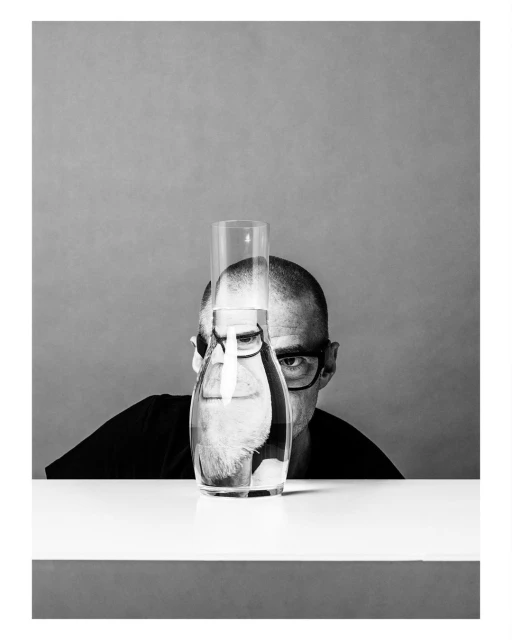 Monochrome portrait of a bald adult man with glasses peering over a table, a water carafe, and a drinking glass with water.