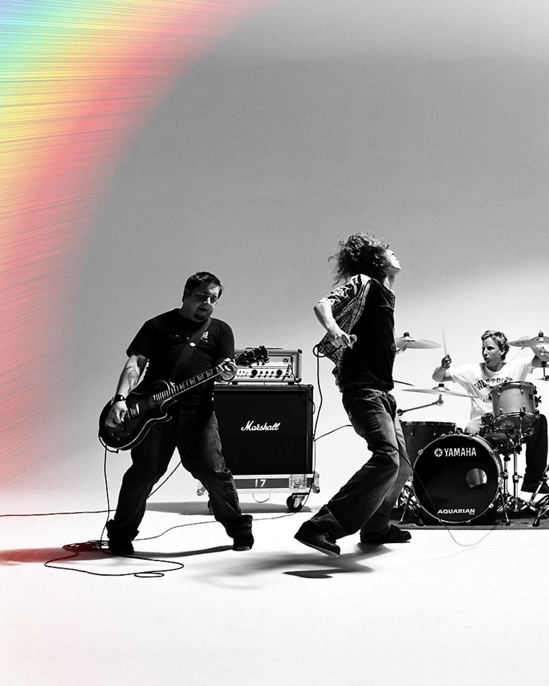 Black and white shot of three musicians performing, guitarist on the left, vocalist in the middle, and a drummer on the right.