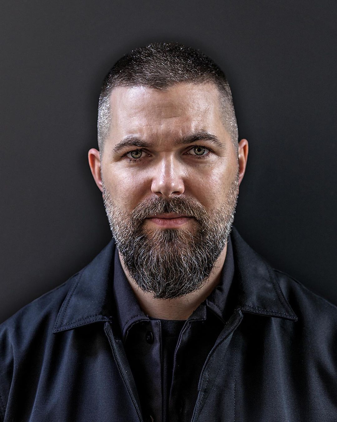 Portrait of an adult male with a beard wearing a dark jacket, looking directly at the camera against a dark grey backdrop.