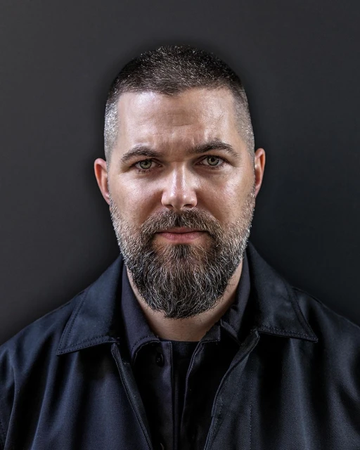 Portrait of an adult male with a beard wearing a dark jacket, looking directly at the camera against a dark grey backdrop.
