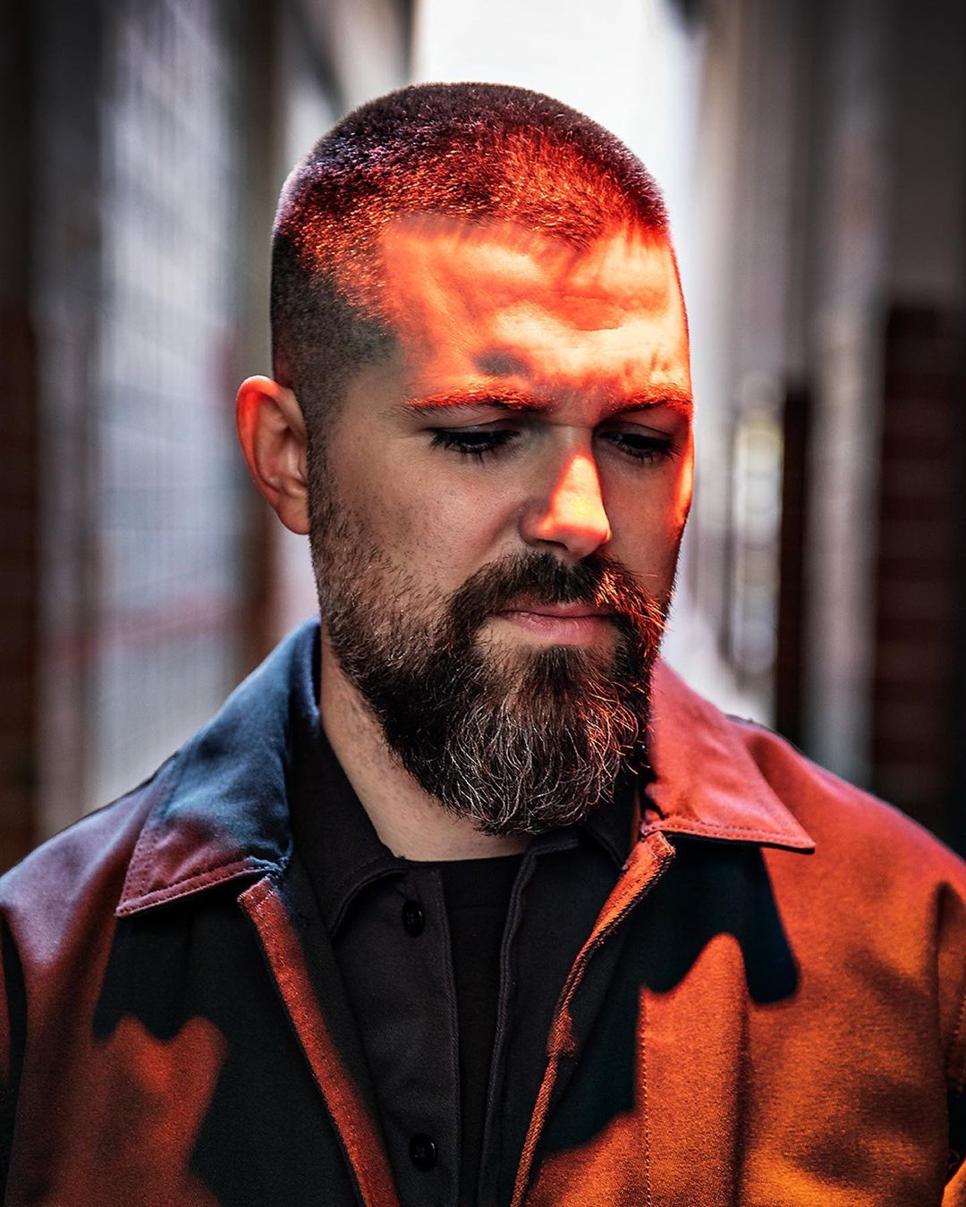 Portrait of a bearded man in a jacket, illuminated by red light against an out-of-focus, textured urban background, looking down.