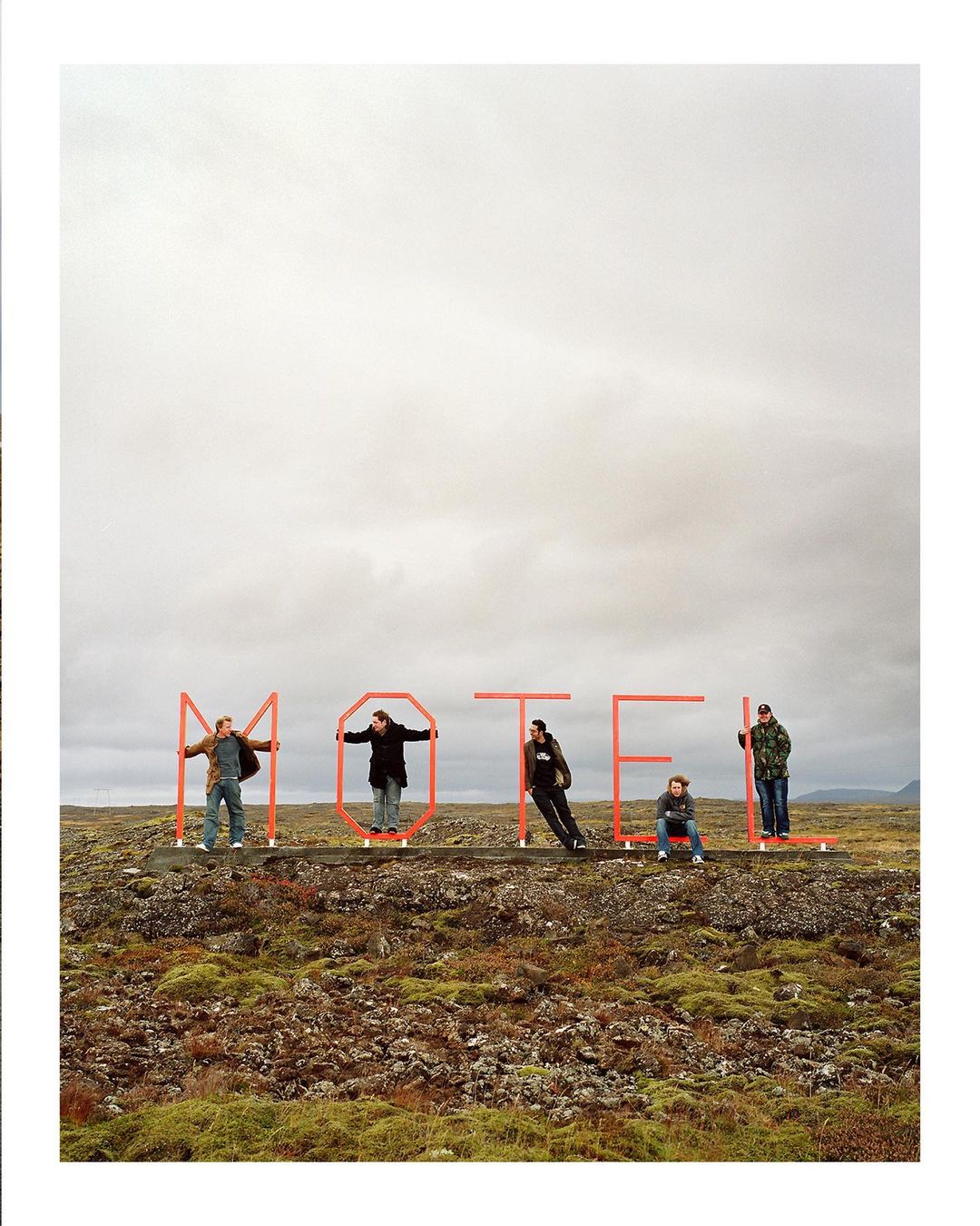 Five people stand behind red metal letters spelling MOTEL in a rocky, mossy landscape under an overcast sky, creating a roadside art scene.