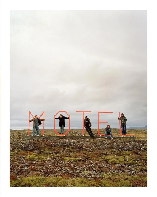 Five people stand behind red metal letters spelling MOTEL in a rocky, mossy landscape under an overcast sky, creating a roadside art scene.