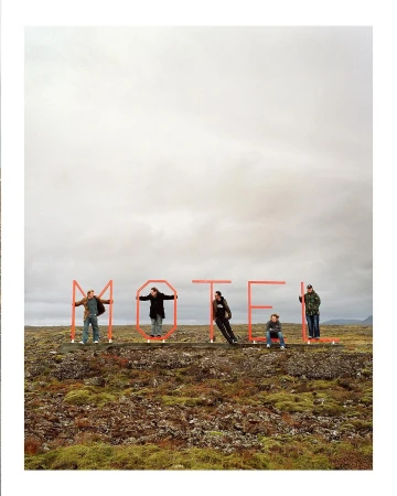 Five people stand behind red metal letters spelling MOTEL in a rocky, mossy landscape under an overcast sky, creating a roadside art scene.