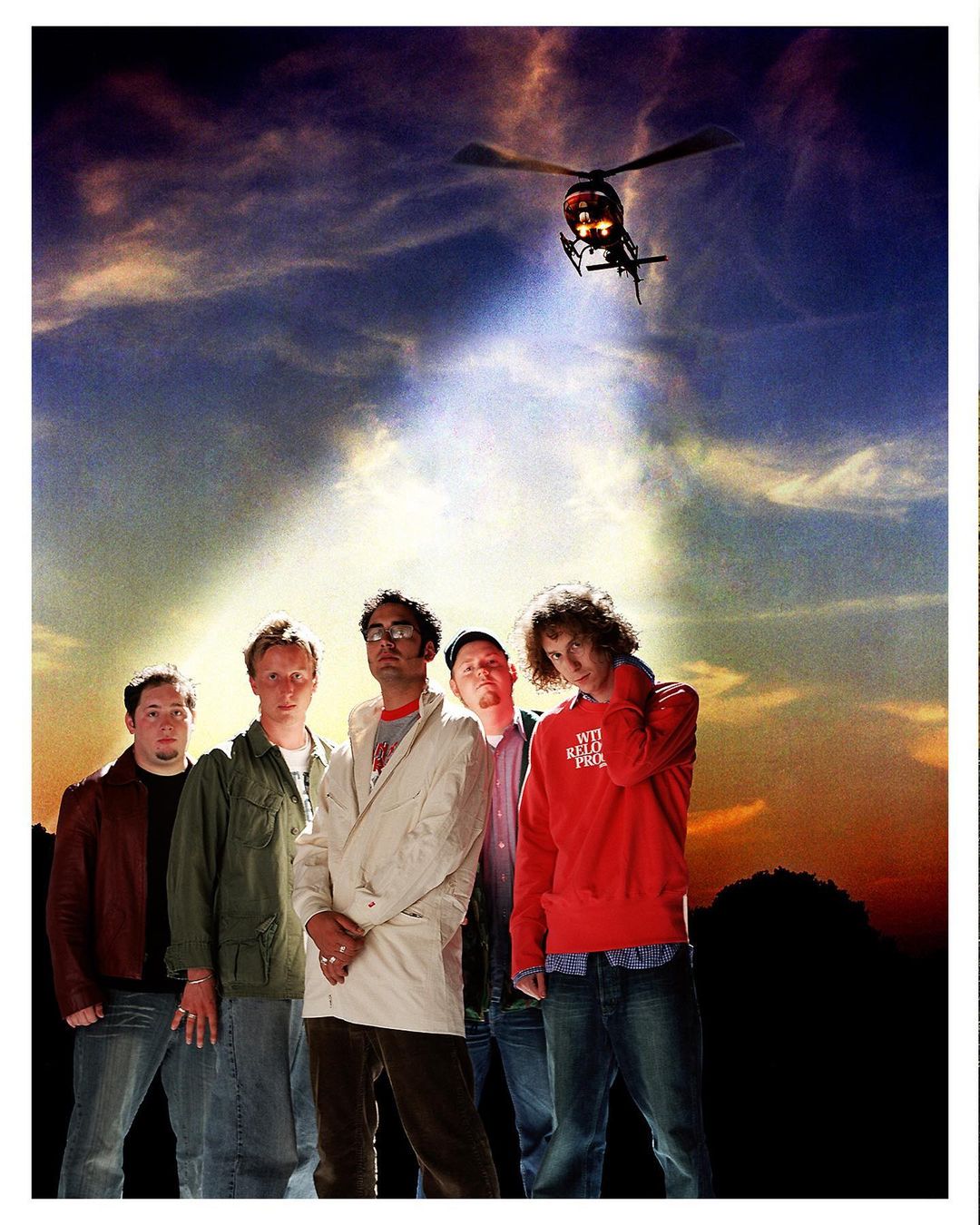 Group portrait of six adults with a helicopter flying overhead against a vibrant cloudy sky at dusk, outdoor setting.