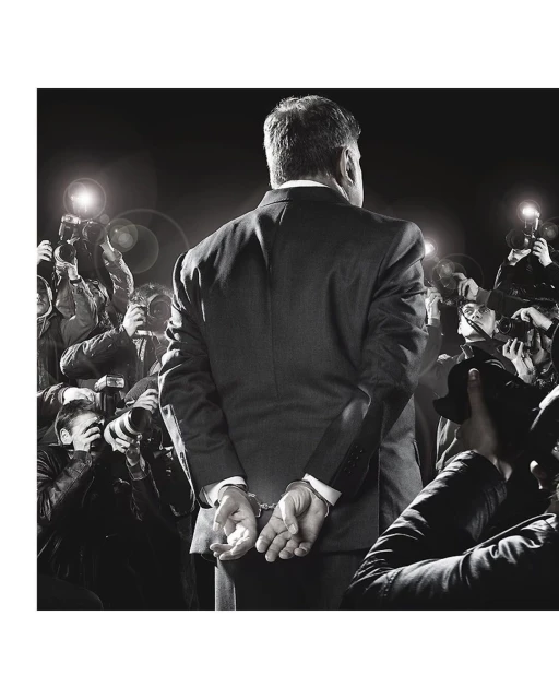 Rear view of an adult male in a suit being escorted by paparazzi and photographers in handcuffs. Dark, monochromatic and formal.