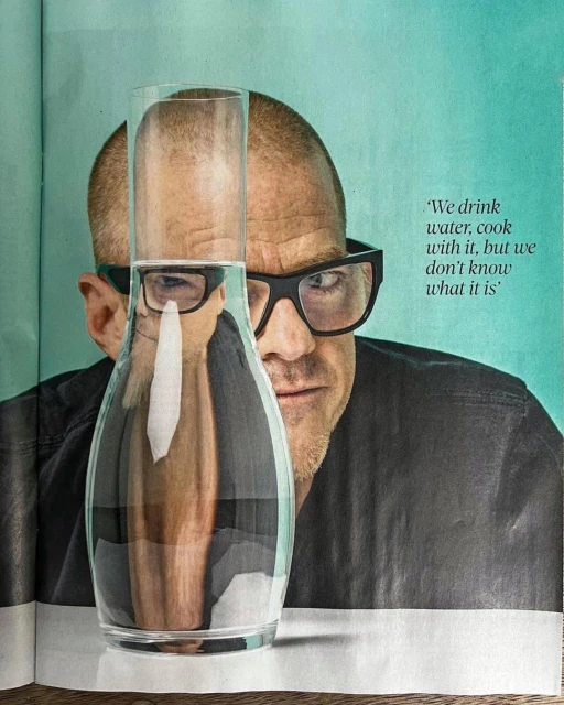 Close-up shot of a bald adult male with glasses peering behind a clear glass pitcher filled with water, in a studio setting with text.