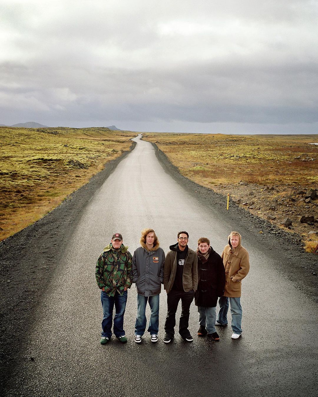 Group of five people stands on rural road, wearing winter clothing, beneath overcast sky, with distant fields and hills.