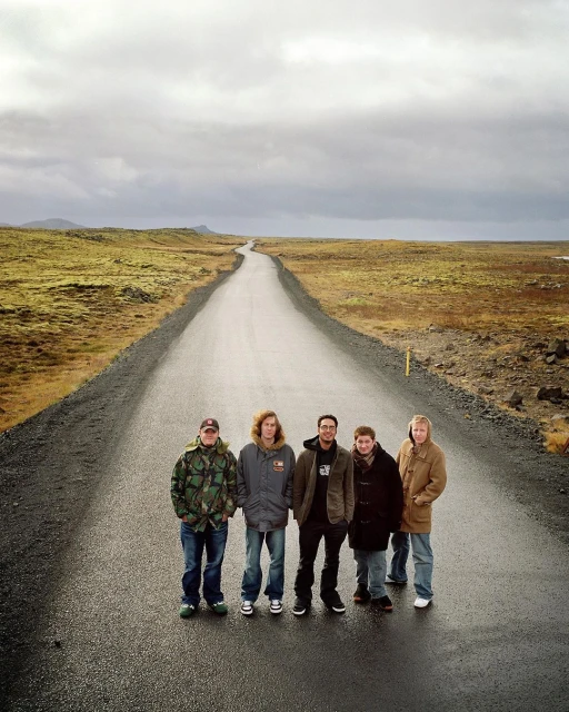 Group of five people stands on rural road, wearing winter clothing, beneath overcast sky, with distant fields and hills.