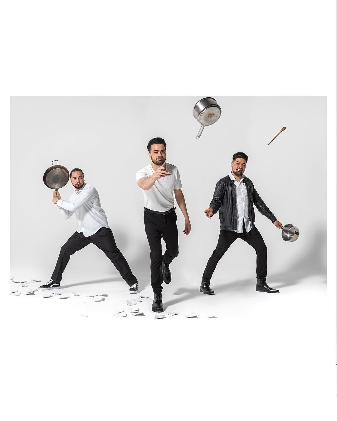 Three men in white shirts and black pants pose with cooking pots and utensils in a studio with a white background and scattered white papers.