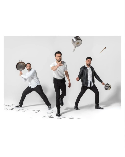 Three men in white shirts and black pants pose with cooking pots and utensils in a studio with a white background and scattered white papers.