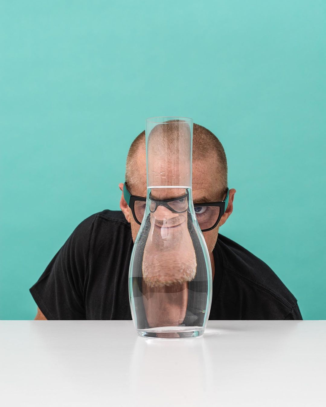 A bald man wearing glasses looks through a carafe of water on a white table against a teal background in a quirky portrait.