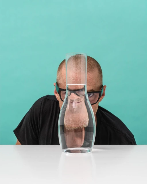 A bald man wearing glasses looks through a carafe of water on a white table against a teal background in a quirky portrait.