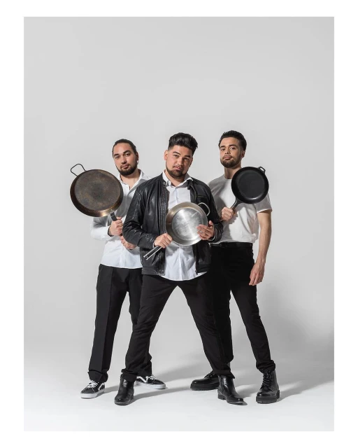 Three men pose indoors with cooking pans, wearing casual outfits and standing against a plain background, studio lighting, medium shot.