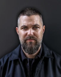 Portrait of a bearded man with short hair wearing a black jacket against a dark background, photographed by Perou.